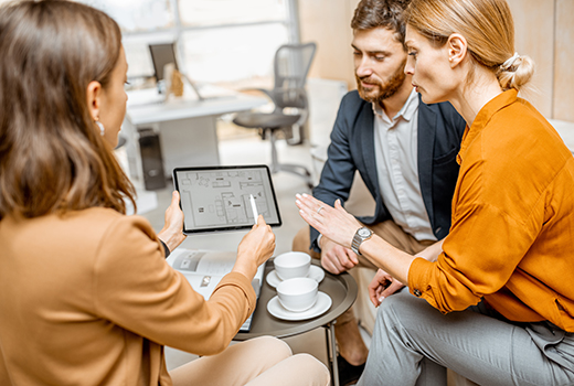 Drei Personen sitzen in einem Büro und betrachten gemeinsam einen Grundriss auf einem Tablet; auf dem Tisch stehen zwei Tassen und eine Untertasse.