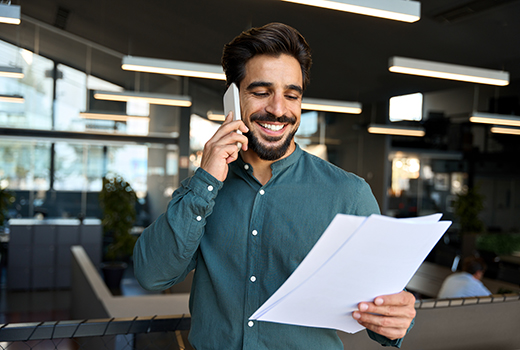 Eine Person steht in einem modernen Büro, hält Papiere in der Hand und spricht am Telefon.